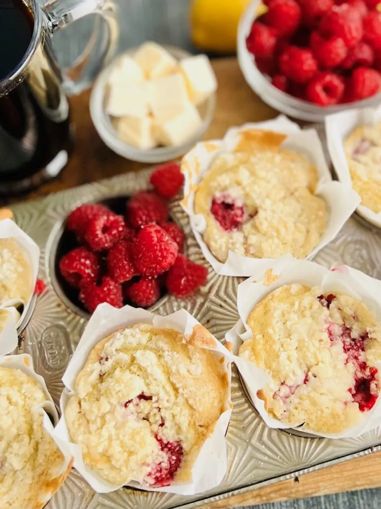 A silver muffin tin holding 5 light colored muffins with speckles of red raspberries popping through. One muffin cavity is filled with bright red raspberries. Next to the muffin tin is a mug of coffee and little cubes of cut up butter.