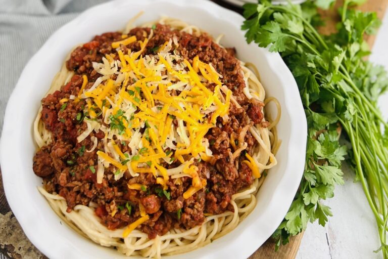 A white, round pie plate with a bottom layer of cooked long spaghetti noodles topped with a red pasta meat sauce. Then on top of that is yellow shredded cheese and chopped up parsley next to fresh bunch of whole parsley.