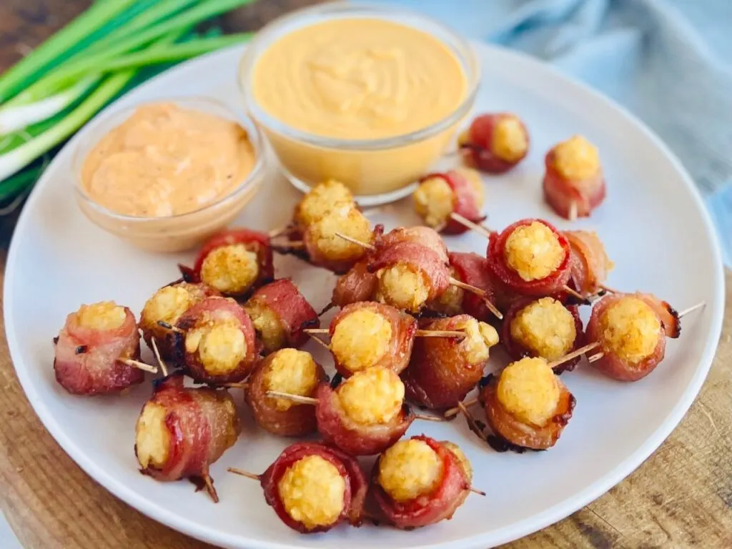 A white serving plate with bacon wrapped tater tots with a toothpick through the middle and 2 dipping sauces nearby.