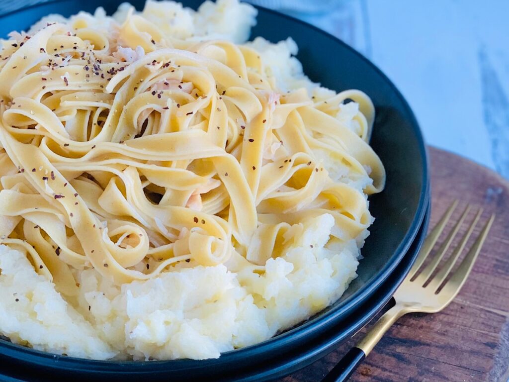 A black bowl filled with a heaping scoop of mashed potatoes and topped with long tender noodles, little chunks of shredded chicken and salt and pepper on top. 
