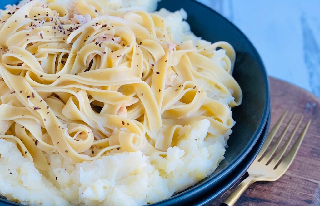 A black bowl filled with a heaping scoop of mashed potatoes and topped with long tender noodles, little chunks of shredded chicken and salt and pepper on top.