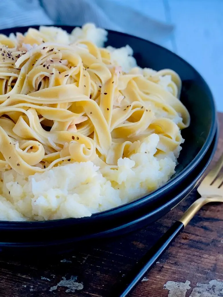 A black bowl filled with a heaping scoop of mashed potatoes and topped with long tender noodles, little chunks of shredded chicken and salt and pepper on top.