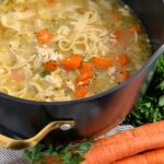 In a large black stock pot there is a mixture of cut carrots, wide noodles, chicken chunks, and broth.