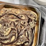 A glass 8x8-inch baking dish filled with a baked marbled brownie mixture with swirls of cream cheese and dessert plates in the background.