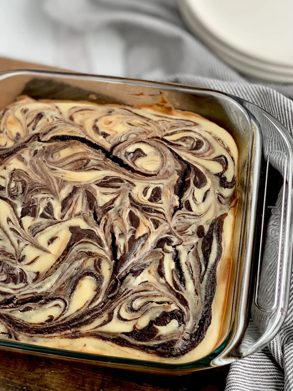 A glass 8x8-inch baking dish filled with a baked marbled brownie mixture with swirls of cream cheese and dessert plates in the background.