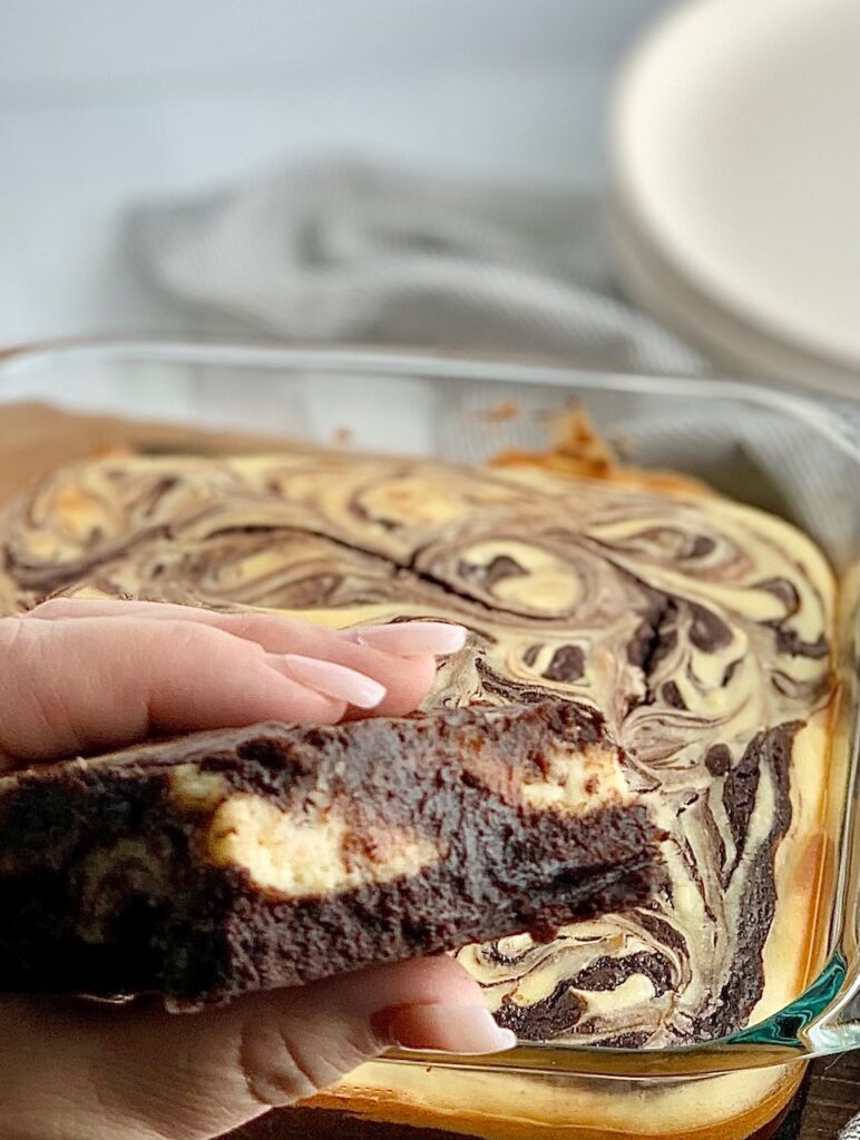 A person folding a cut out brownie with chunks of white cream cheese in it with an 8x8-inch baking dish filled with a baked marbled brownie mixture with swirls of cream cheese and dessert plates in the background.