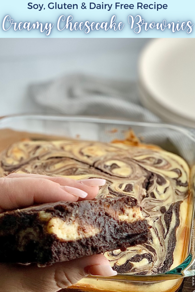 A person folding a cut out brownie with chunks of white cream cheese in it with an 8x8-inch baking dish filled with a baked marbled brownie mixture with swirls of cream cheese and dessert plates in the background.