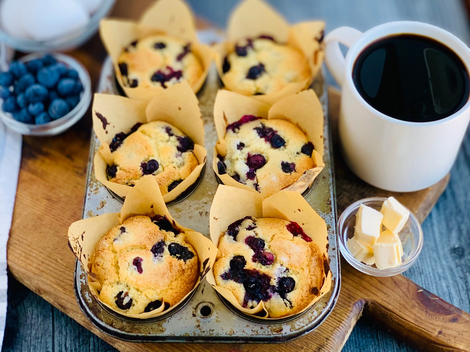 A cutting board with a 6 cavity muffin tin filled with light brown muffin s filled with loads of blueberries.