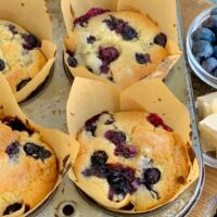 A 6 cavity metal muffin pan filled with light brown tulip cupcake liners. Inside the liners is a soft brown colored muffin with blueberries everywhere.