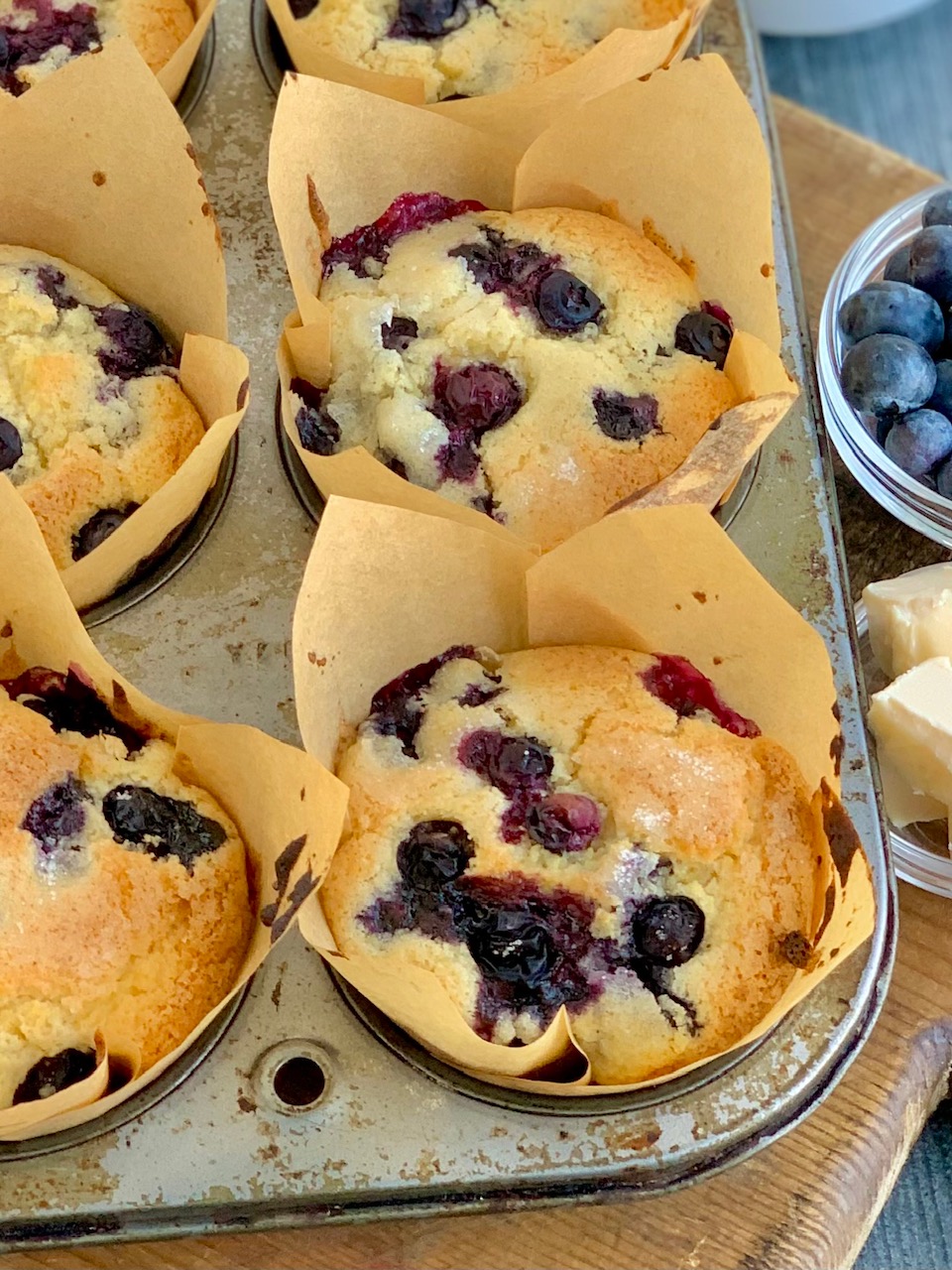 A 6 cavity metal muffin pan filled with light brown tulip cupcake liners. Inside the liners is a soft brown colored muffin with blueberries everywhere next to a bowl of fresh blueberries and butter.