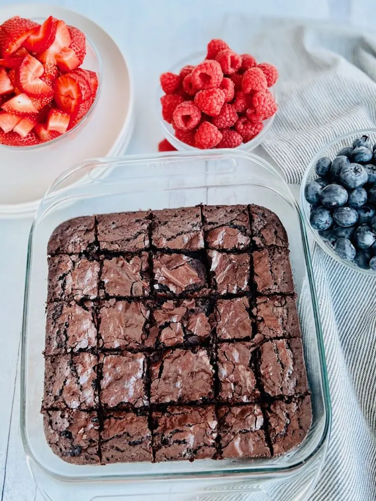 A small bowl of cut strawberries, fresh red raspberries, another bowl of raspberries, and an 8x8-inch pan of cut brownies.