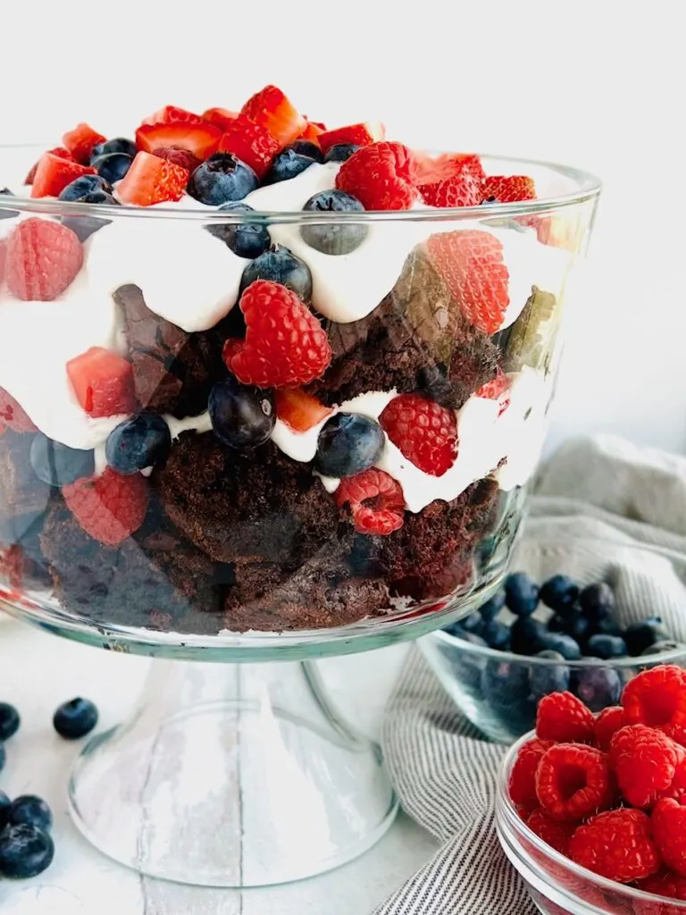 A large glass bowl filled with layers of small brownie squares, cut fresh fruit like blueberries, raspberries, and strawberries, and a creamy, white cocowhip.