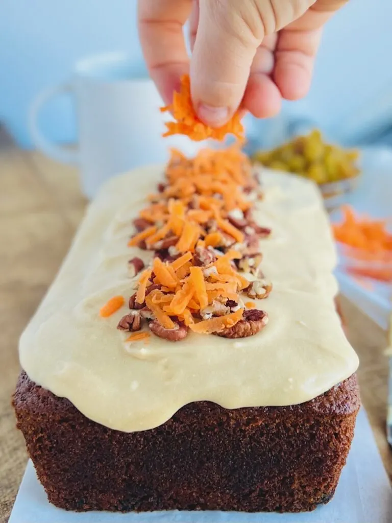 A freshly baked loaf of dark carrot cake loaf with a light brown cream cheese frosting over the top and chopped walnuts. A hand is shown sprinkling fresh shredded carrots over the top of the walnuts. 