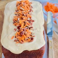 A freshly baked loaf of dark carrot cake loaf with a light brown cream cheese frosting over the top and chopped walnut and freshly shredded carrots down the middle.
