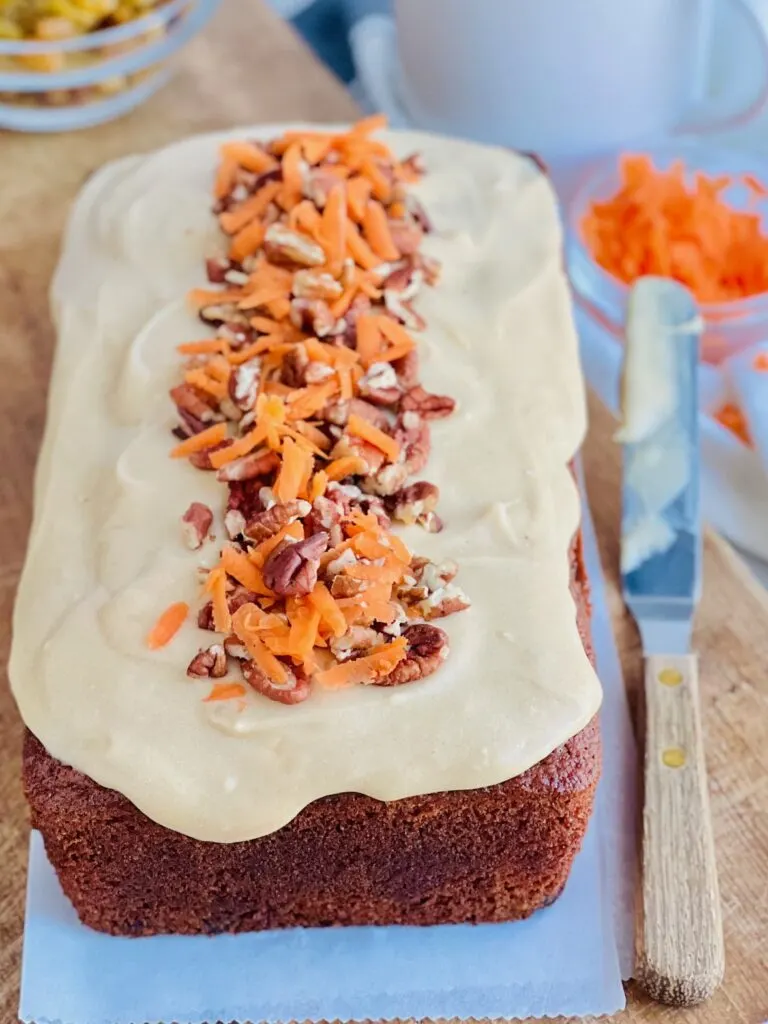 A freshly baked loaf of dark carrot cake loaf with a light brown cream cheese frosting over the top and chopped walnut and freshly shredded carrots down the middle next to a small bowl of shredded carrots.