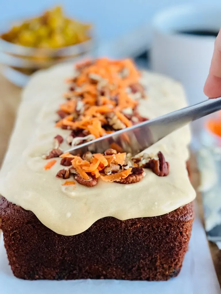 A freshly baked loaf of dark carrot cake loaf with a light brown cream cheese frosting over the top and chopped walnuts and freshly shredded carrots down the middle. A knife is cutting into the bread.