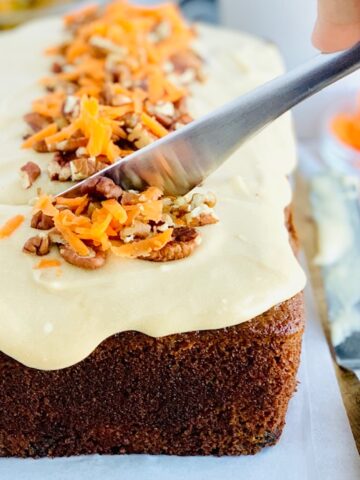 A knife is cutting into a 9x5-inch loaf of dark colored carrot cake loaf. The cake bread is topped with a warm colored brown sugar cream cheese frosting with chopped walnuts and shredded carrots down the middle.