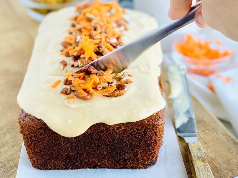 A knife is cutting into a 9x5-inch loaf of dark colored carrot cake loaf. The cake bread is topped with a warm colored brown sugar cream cheese frosting with chopped walnuts and shredded carrots down the middle.