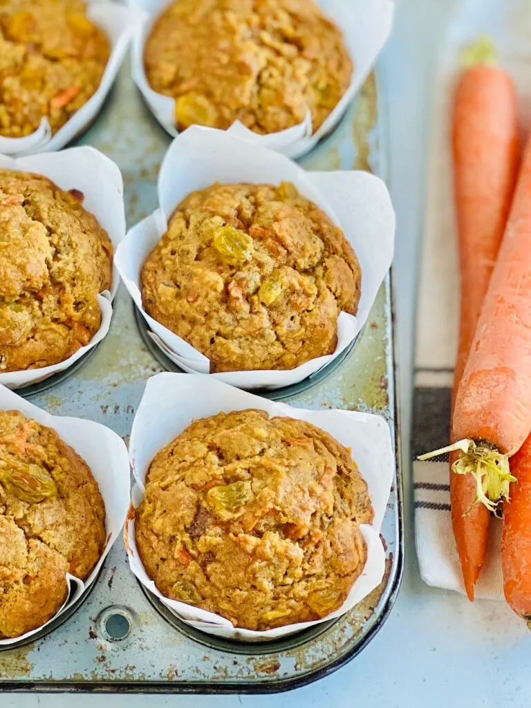 A 6 cavity muffin tin filled with brown and orange colored muffins with golden raisins showing through next to 3 large carrots on a table. 