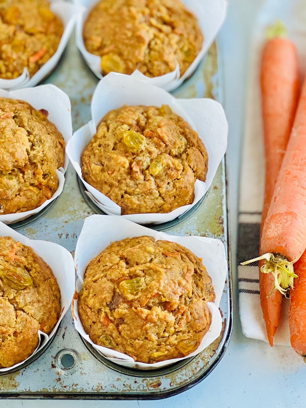 A 6 cavity muffin tin filled with brown and orange colored muffins with golden raisins showing through next to 3 large carrots on a table. with