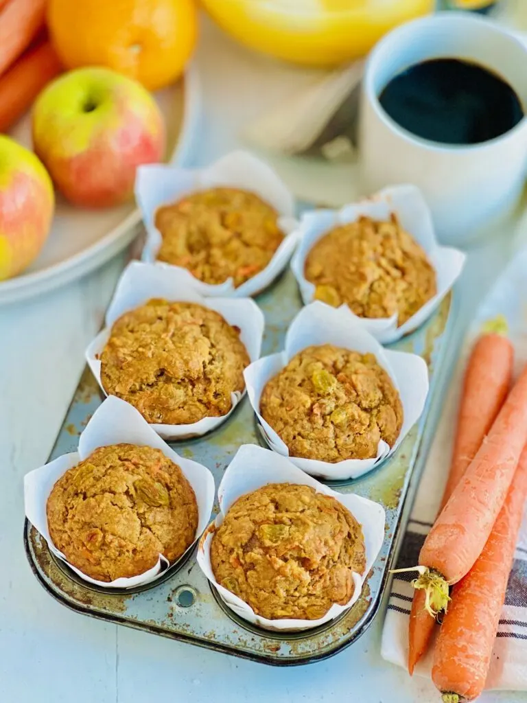 A 6 cavity muffin tin filled with brown and orange colored muffins with golden raisins showing through next to 3 large carrots on a table with a mug of coffee.