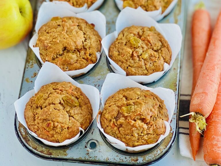 4 brown orange colored muffins in a muffin tin next to a shiny apple and 3 large carrots.