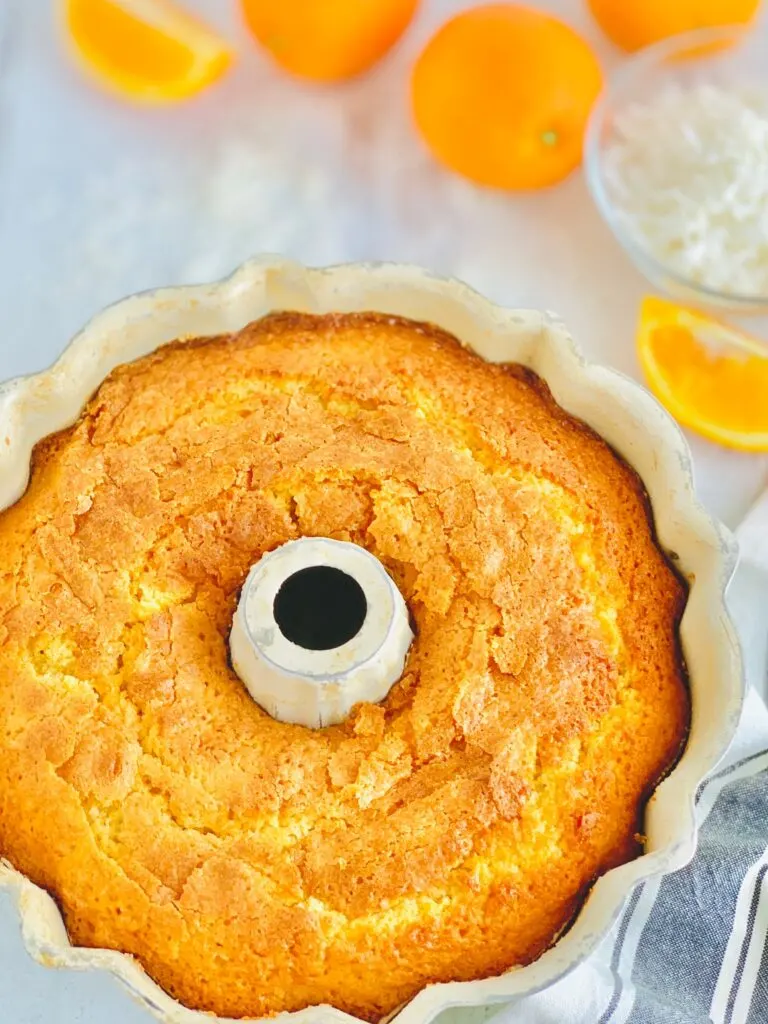 A beautiful orange colored baked cake in a bundt pan with slices oranges in the back ground.