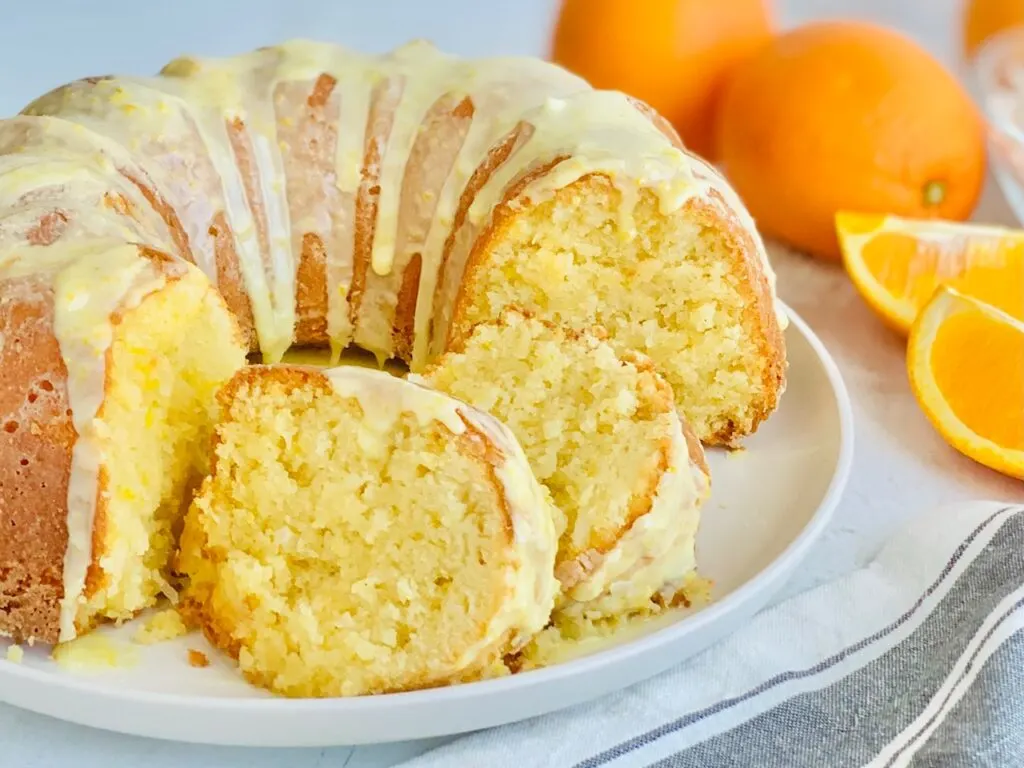 A circle shaped bundt cake with shredded coconut and fresh orange zest cut into slices. There is a light colored glaze over the top of the cake.