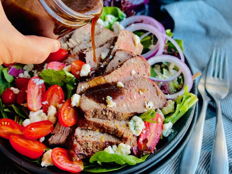 A large dinner salad bowl filled with salad greens, sliced cherry tomatoes, cut purple onion, dairy free bleu cheese crumbles, and thin slices of grilled steak with a dark dressing being poured over it.
