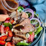 A large black plate layered with colorful salad greens, thin strips of purple onion, cherry tomatoes cut in half, blue cheese crumbles and a hand pouring a dressing over thin slices of steak.