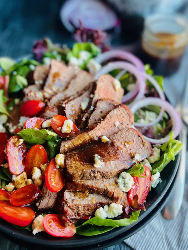 A large black plate layered with colorful salad greens, thin strips of purple onion, cherry tomatoes cut in half, blue cheese crumbles and a hand pouring a dressing over thin slices of steak. 