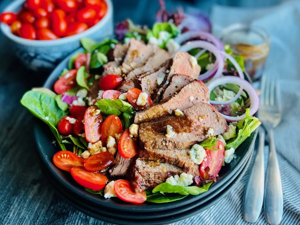 A large black plate layered with colorful salad greens, thin strips of purple onion, cherry tomatoes cut in half, blue cheese crumbles and dressing over thin slices of steak. 