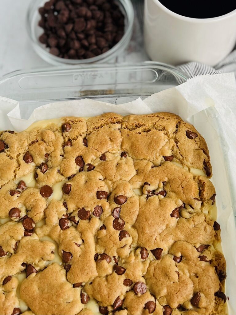 A small glass pan of baked chocolate chip cookie with little sneak peaks of a white cream cheese layer.