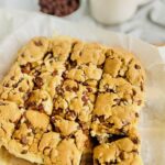 A square of chocolate chip cookie batter baked into bars not cookies. There is a bottom layer of baked chocolate chip cookie then a layer of white cream cheese and another layer of chocolate chip cookie on top. The square is cut into bars.