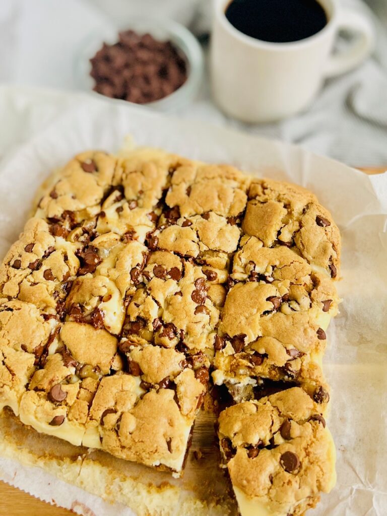 A square of chocolate chip cookie batter baked into bars not cookies. There is a bottom layer of baked chocolate chip cookie then a layer of white cream cheese and another layer of chocolate chip cookie on top. The square is cut into bars.