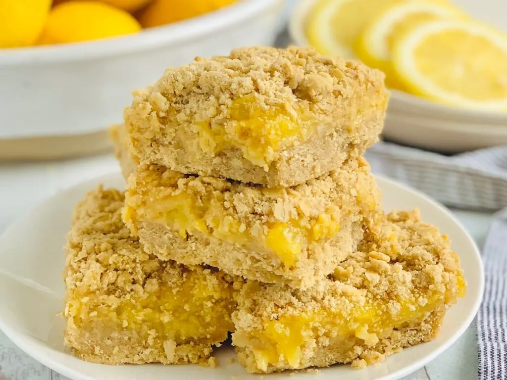 A white plate filled with cut dessert square. The first layer of the bar is a light brown color followed by a gooey yellow curd then topped with another light brown oat layer.