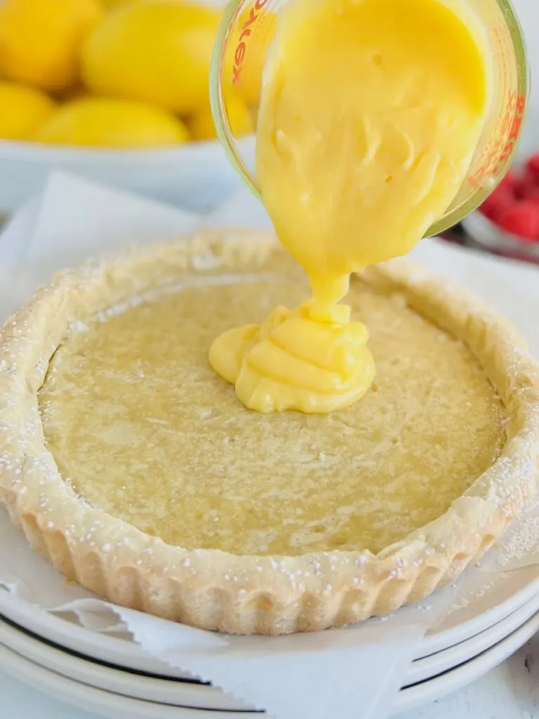 A white tart crust with yellow lemon curd being poured into the tart crust.