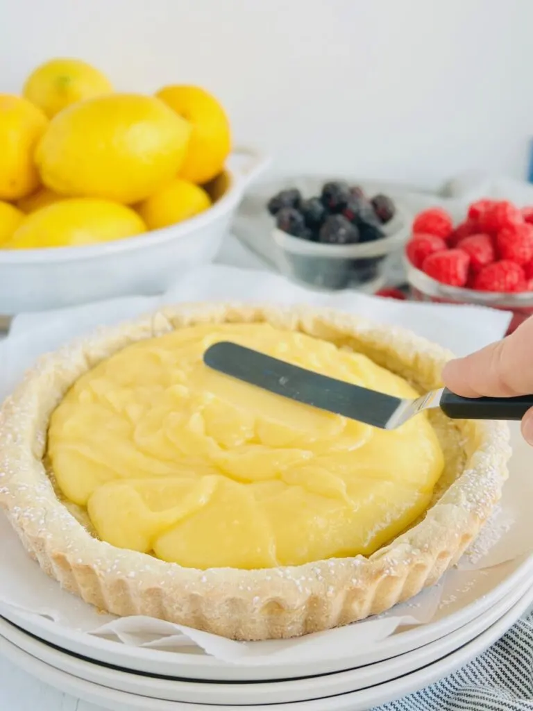 A white tart crust with a yellow, lemon curd filling being evenly filled in the tart with an angled spatula.