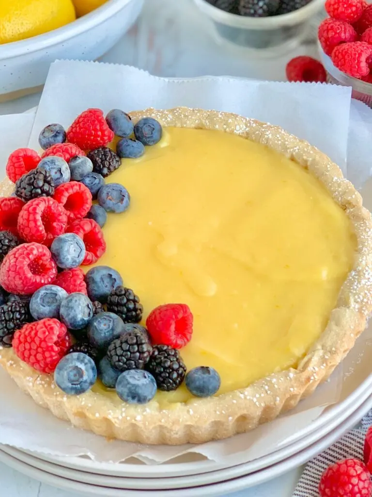 A white tart crust with a yellow, lemon curd filling topped on one side with blueberries, blackberries, and raspberries.