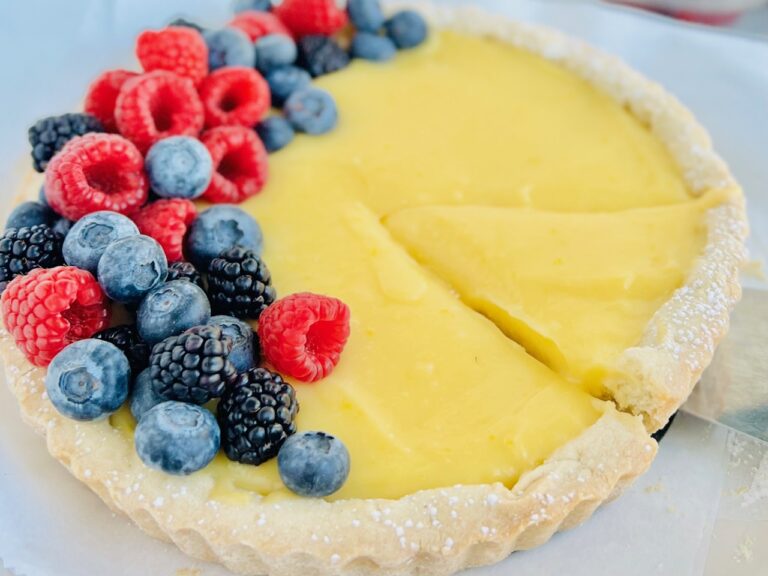A white tart with yellow lemon curd filling inside and topped with fruit. A piece is cut and being removed.