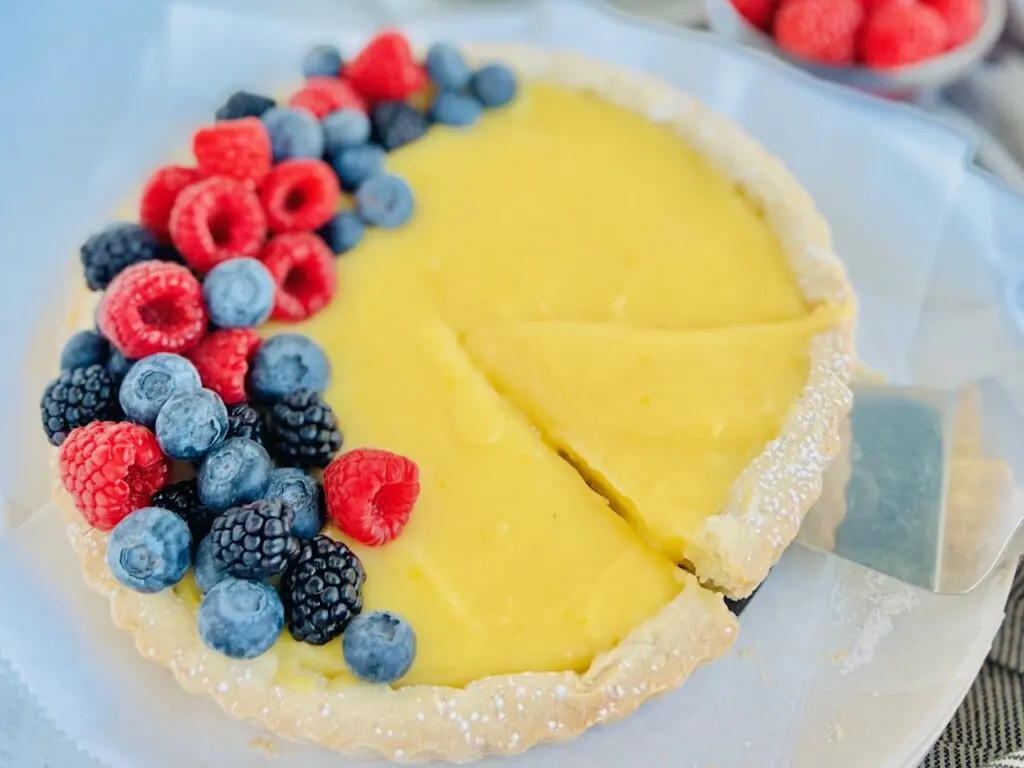 A white tart with yellow lemon curd filling inside and topped with fruit. A piece is cut and being removed.