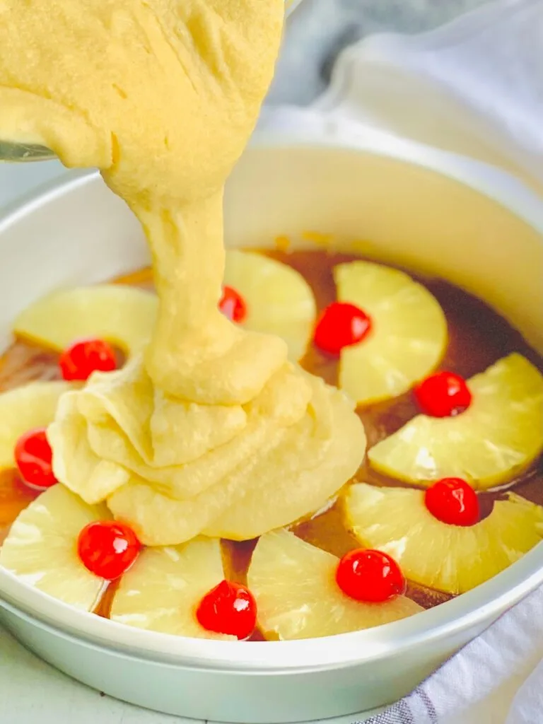 Yellow cake batter is being poured over cherries, pineapple slices, and the brown sugar mixture.