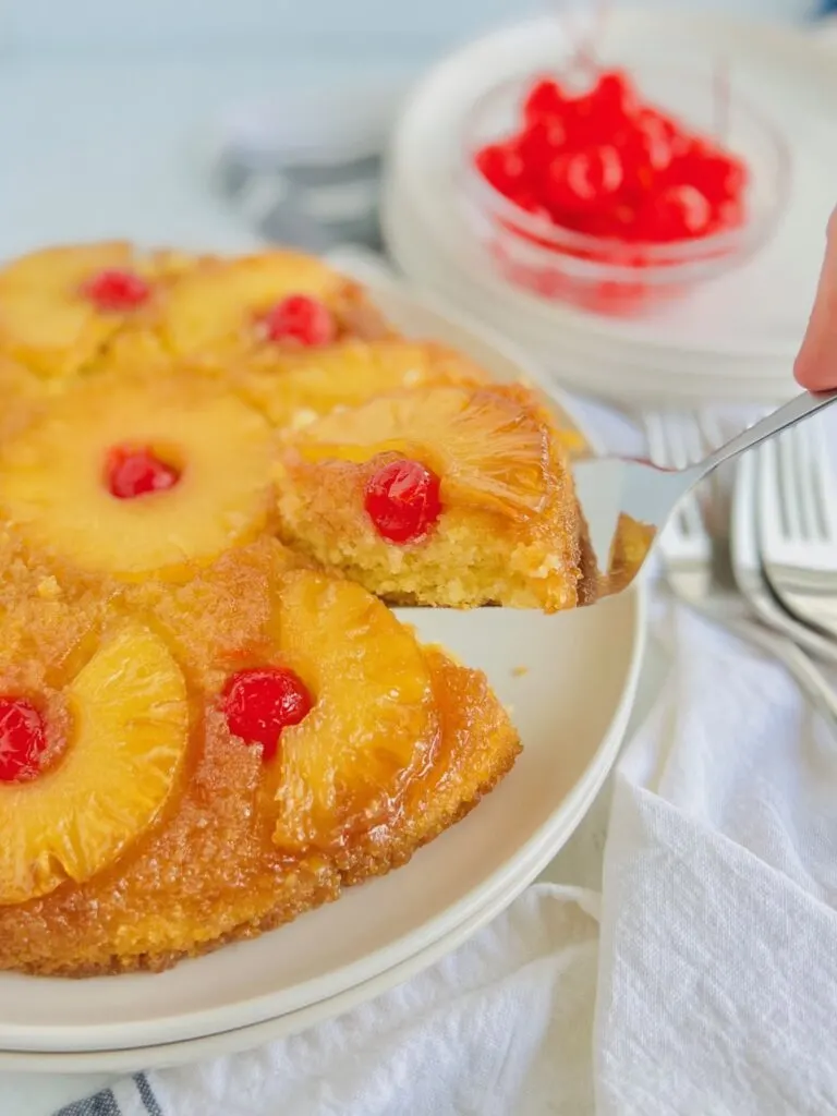 A round yellow brown cake with half pineapple slices around the the sides and a red Cherrie in the middle in the middle of the curved pineapple slice. The cake has a slice cut out of it and a serving spatula is removing the slice.