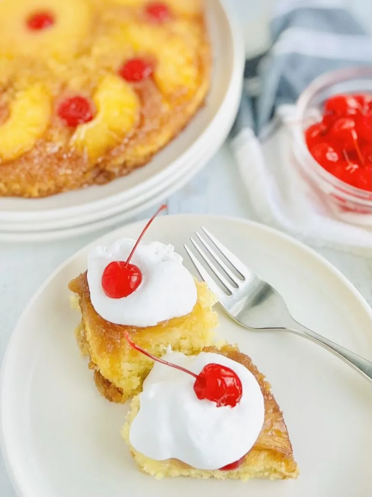 A round yellow brown cake with half pineapple slices around the the sides and a red Cherrie in the middle in the middle of the curved pineapple slice. There is a white dessert plate with 2 slices of cake on it with white whipped topping and cherries near a fork.