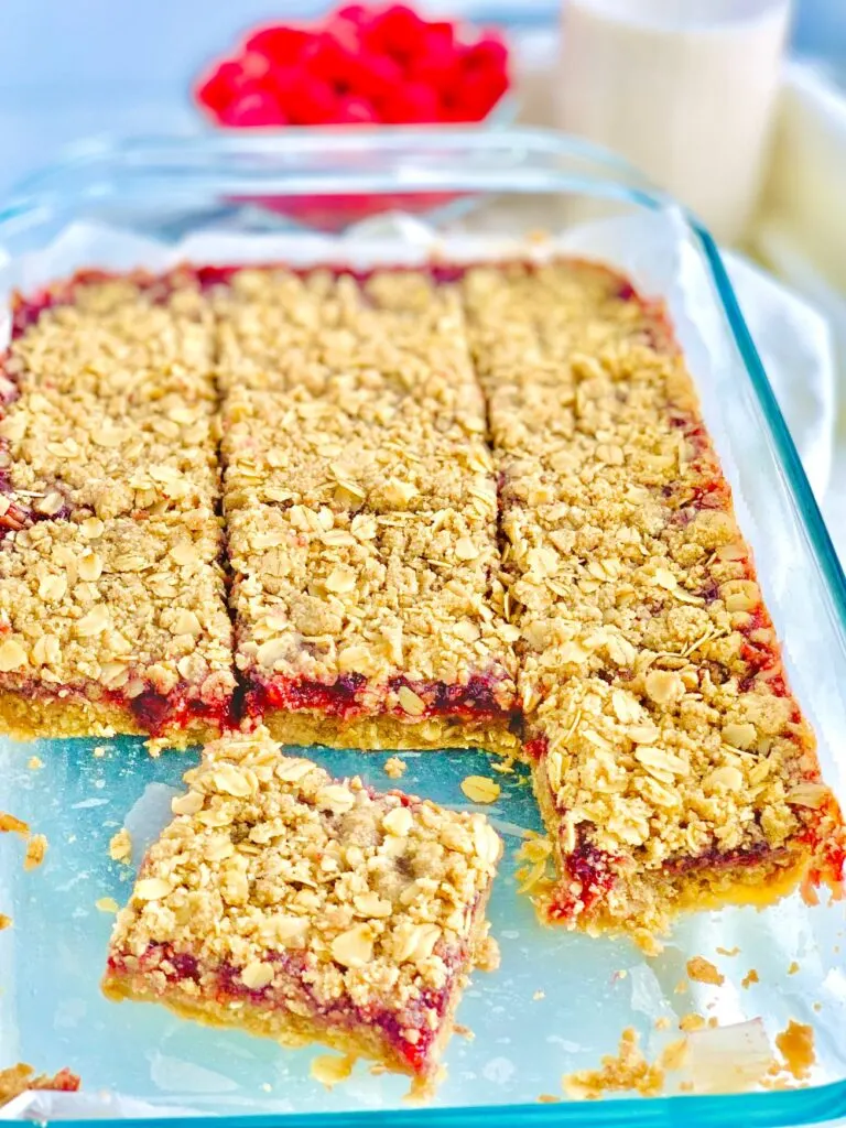 A glass 9x13-inch baking dish filled with a layer of oatmeal crumble, then a red raspberry layer and lastly it is topped with another layer of oatmeal crumble. The bars have been cut into large square slices.