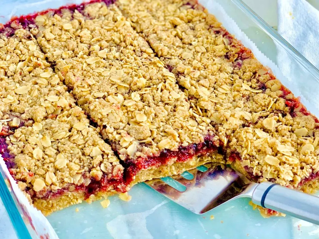 A glass 9x13-inch baking dish filled with a layer of oatmeal crumble, then a red raspberry layer and lastly it is topped with another layer of oatmeal crumble. The bars have been cut into large square slices.