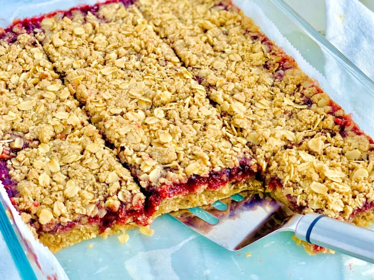 A glass baking dish with a layer of oatmeal crumble, raspberry preserves, and another layer of oatmeal crumble cut into squares and a serving spatula removing some.