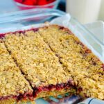 A glass 9x13-inch baking dish filled with a layer of oatmeal crumble, then a red raspberry layer and lastly it is topped with another layer of oatmeal crumble. The bars have been cut into large square slices.