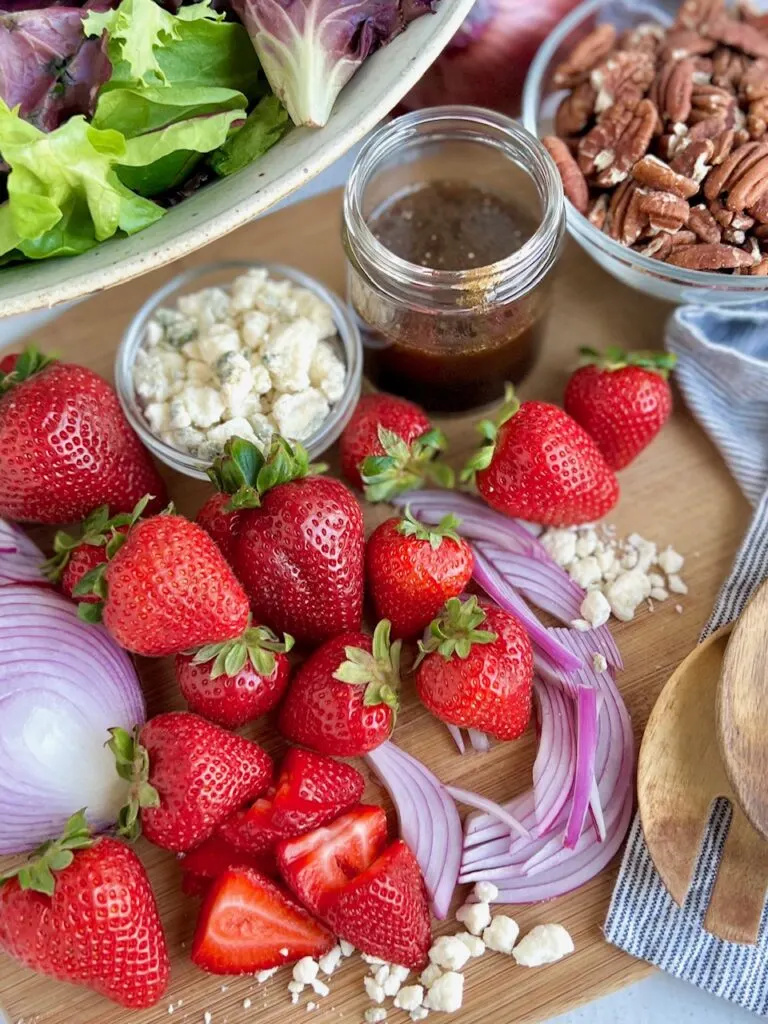 A cutting board filled with salad ingredients like slices red onions, strawberries, a little bowl of dairy free bleu cheese, a bowl of toasted pecans, and a glass jar of brown dressing.