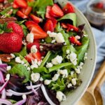 A large bowl heaping with green and purple salad mix, sliced bright red strawberries, brown pecans, thin slices of red onions, and white crumbles of dairy free bleu cheese.